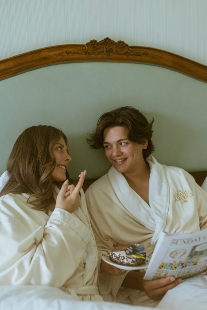 Blake and Kaylee share a bite of anniversary cake in bed, leaning into the whimsical, French-Sunday-morning mood they envisioned for their session at Grand America.