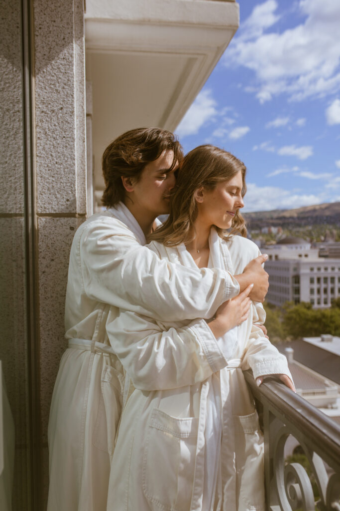 Couple in matching white Grand America robes embracing on hotel balcony overlooking Salt Lake City