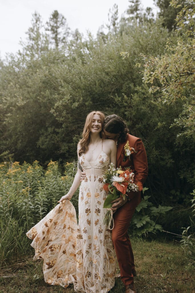 Bride embraces groom from behind as they stand in a quiet forest clearing on their wedding day